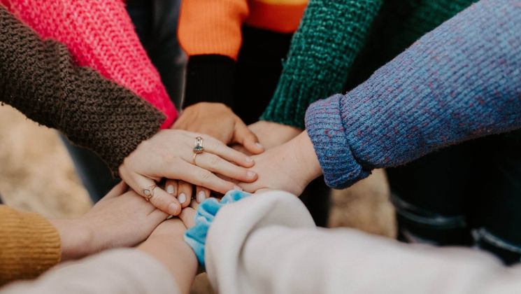 Verschiedene Hände, die übereinandergelegt sind. Die Personen tragen bunte Pullover. Symbol für Gemeinschaft und Unterstützung.
