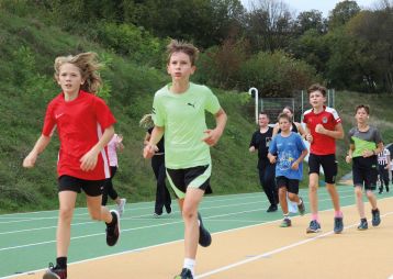 Eine Gruppe von Kindern joggt auf einer Leichtathletikanlage. Es sind verschiedene Sportkleidungen zu sehen und die Kinder wirken motiviert.
