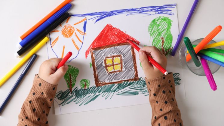 Ein Kind zeichnet ein farbenfrohes Haus mit verschiedenen Markern auf einem Blatt Papier.