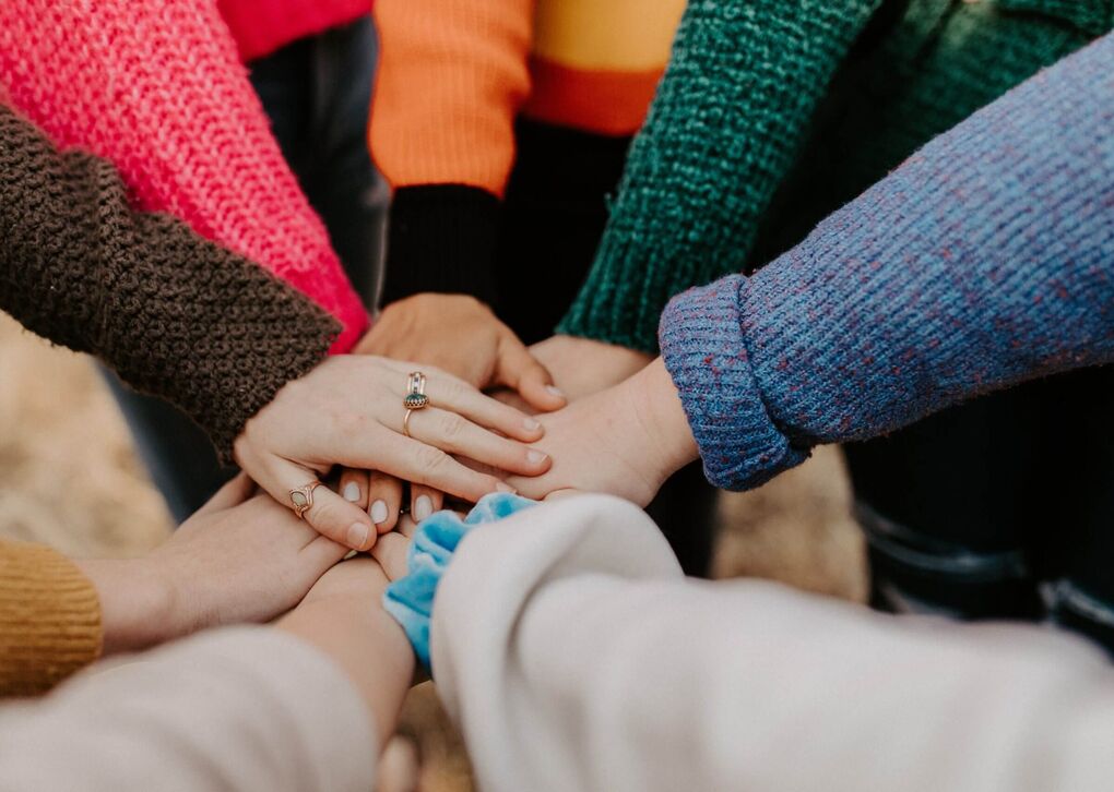 Verschiedene Hände, die übereinandergelegt sind. Die Personen tragen bunte Pullover. Symbol für Gemeinschaft und Unterstützung.