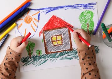 Ein Kind zeichnet ein farbenfrohes Haus mit verschiedenen Markern auf einem Blatt Papier.
