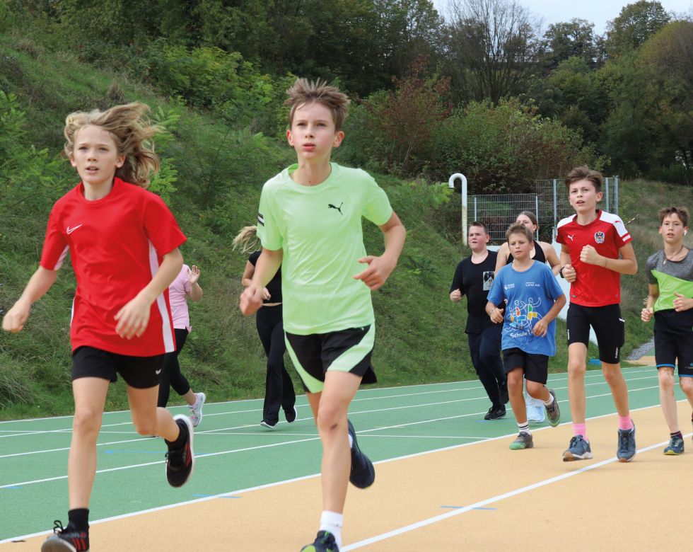 Eine Gruppe von Kindern joggt auf einer Leichtathletikanlage. Es sind verschiedene Sportkleidungen zu sehen und die Kinder wirken motiviert.