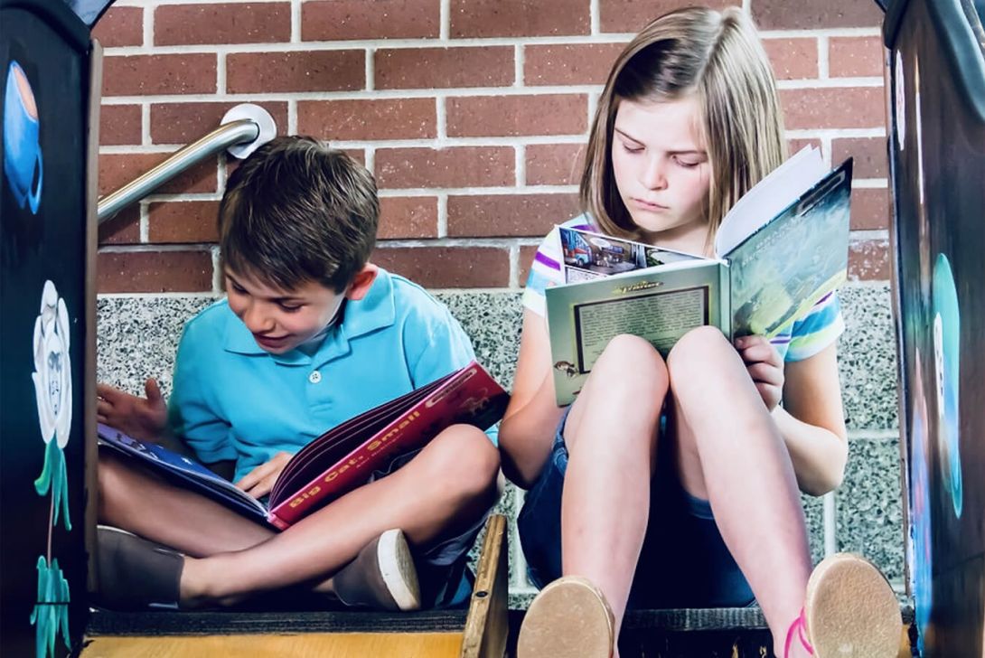 Zwei Kinder sitzen auf einer Rutsche und lesen Bücher, eines lacht und zeigt auf die Seite, während das andere ernsthaft liest.