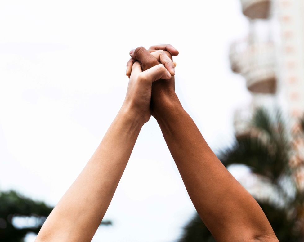 Zwei Hände halten sich hoch, symbolisieren Einheit und Solidarität. Im Hintergrund sind unscharfe Pflanzen und Gebäude sichtbar.