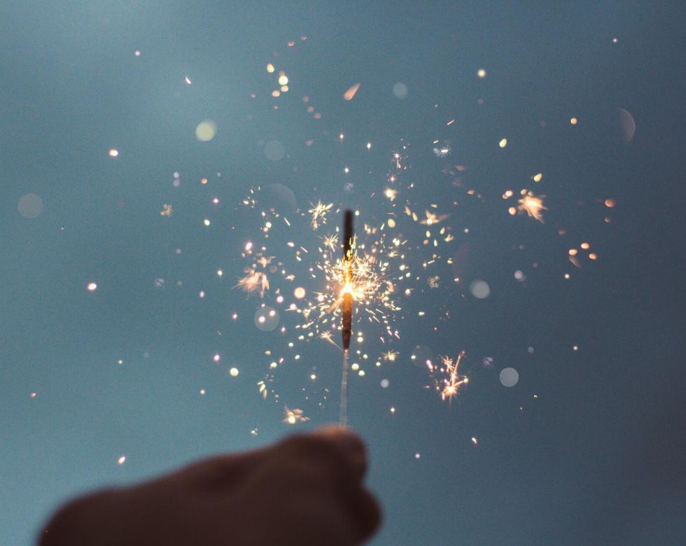 Eine Hand hält ein Wunderkerze, die funkelnde Lichter in den Himmel ausstrahlt. Der Hintergrund ist in sanften Blautönen gehalten.