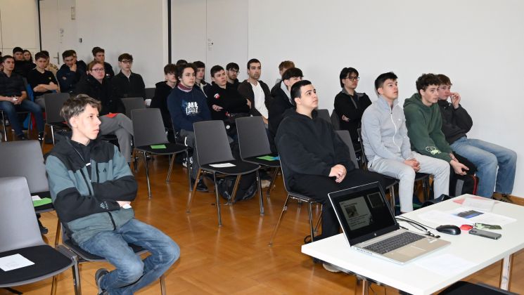 Ein Dozent spricht vor einer Gruppe von Schülern in einem Klassenzimmer. Die Teilnehmer hören aufmerksam zu und nehmen Notizen.