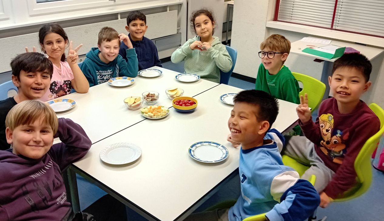 Eine Gruppe fröhlicher Kinder sitzt um einen Tisch mit verschiedenen Snacks. Sie lächeln und haben Spaß miteinander.