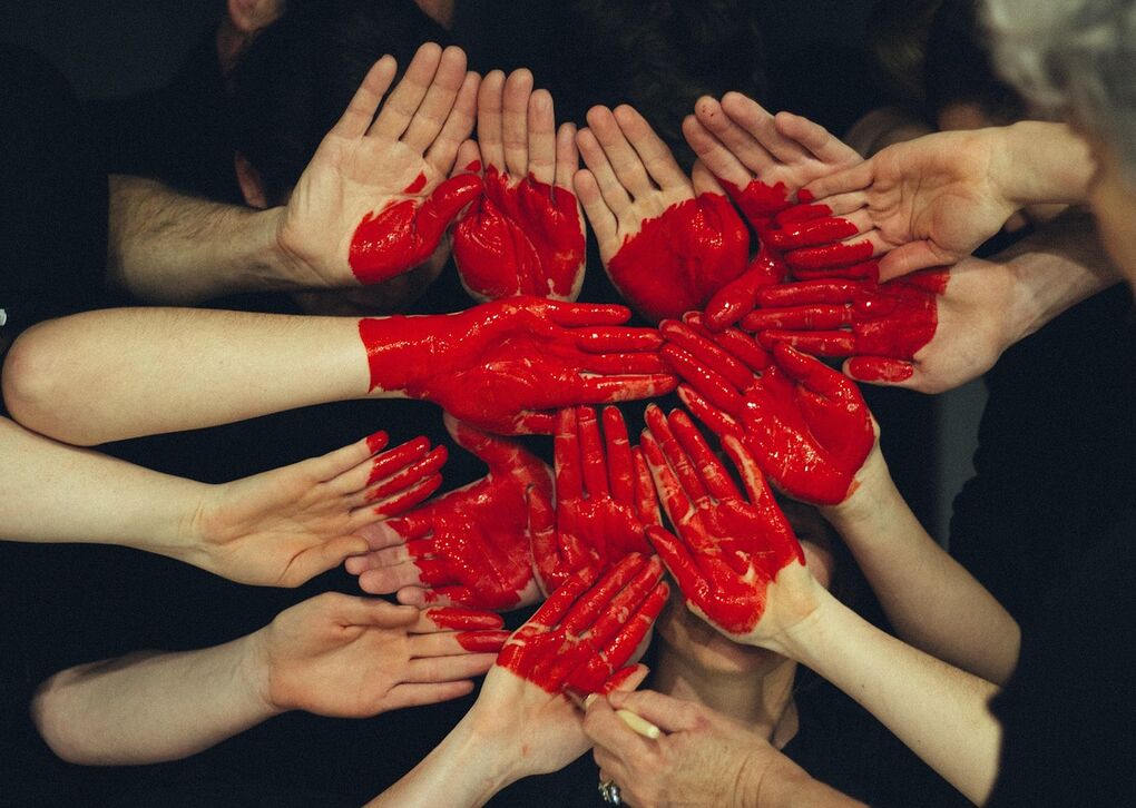 Eine Gruppe von Händen, einige mit roter Farbe bemalt, bildet ein Herz-Muster. Ausdruck von Gemeinschaft und Kreativität.