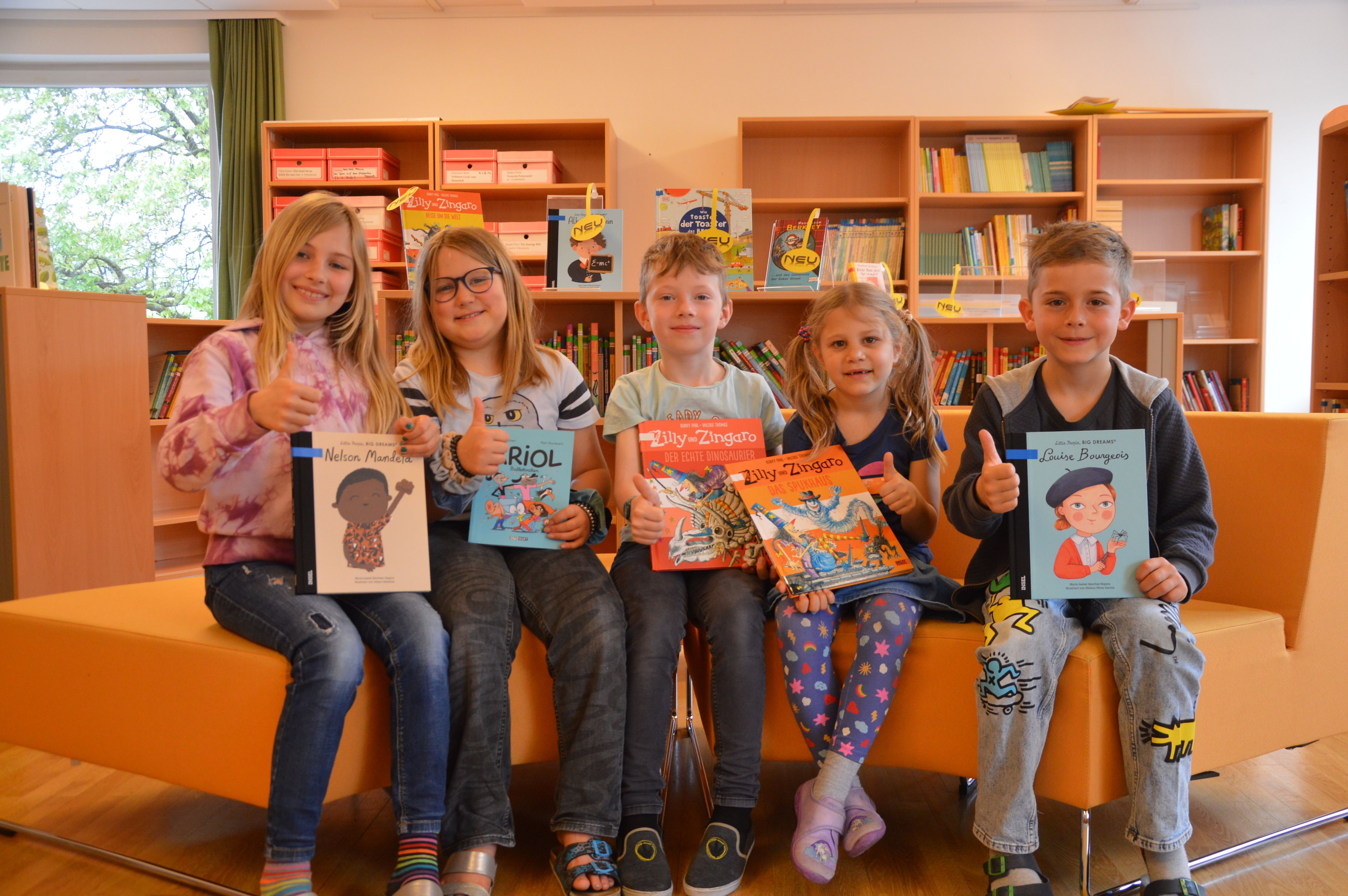 Fünf Kinder lächeln und zeigen Daumen hoch, während sie ihre Lieblingsbücher in einer Bibliothek halten.