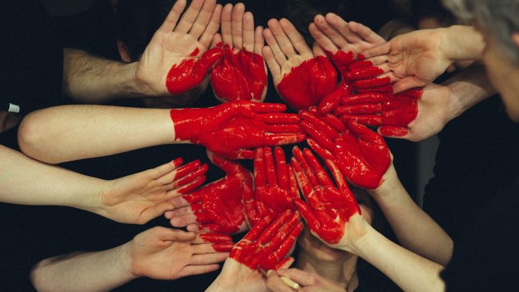 Eine Gruppe von Händen, einige mit roter Farbe bemalt, bildet ein Herz-Muster. Ausdruck von Gemeinschaft und Kreativität.