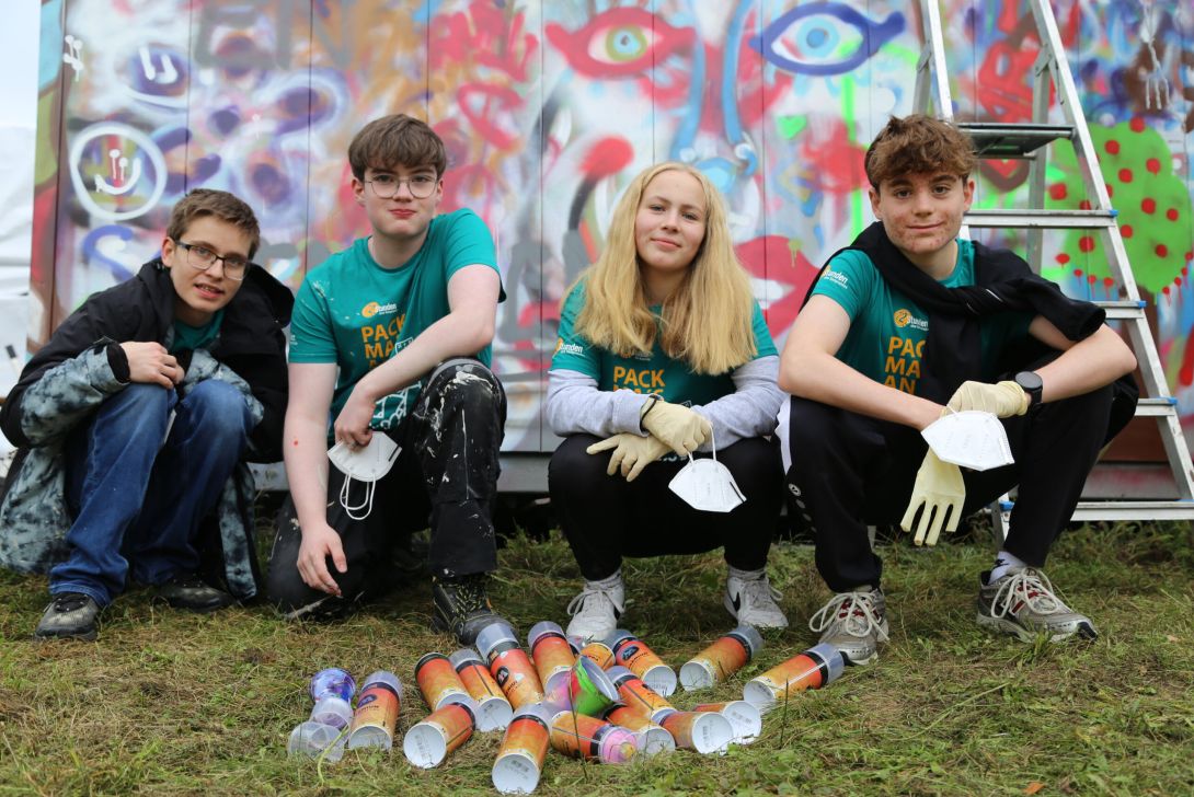 Eine Gruppe von vier jungen Menschen sitzt auf dem Gras vor einer bunten Graffiti-Wand und hält Masken in den Händen.