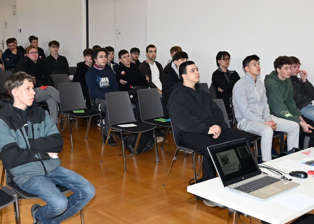Ein Dozent spricht vor einer Gruppe von Schülern in einem Klassenzimmer. Die Teilnehmer hören aufmerksam zu und nehmen Notizen.