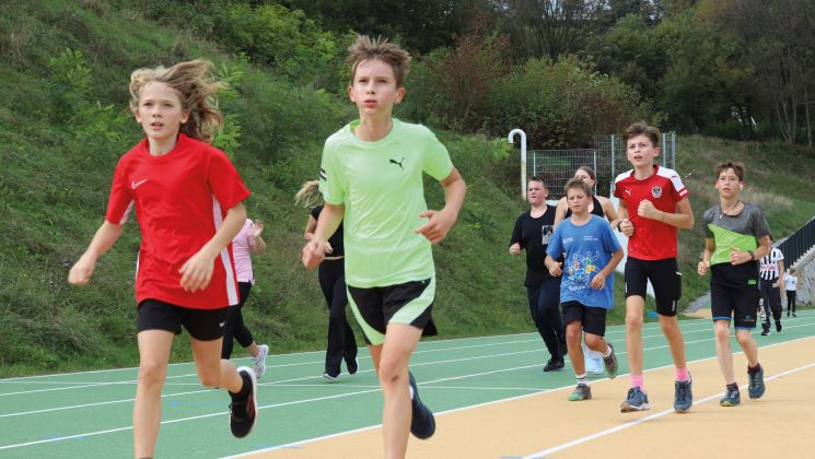 Eine Gruppe von Kindern joggt auf einer Leichtathletikanlage. Es sind verschiedene Sportkleidungen zu sehen und die Kinder wirken motiviert.