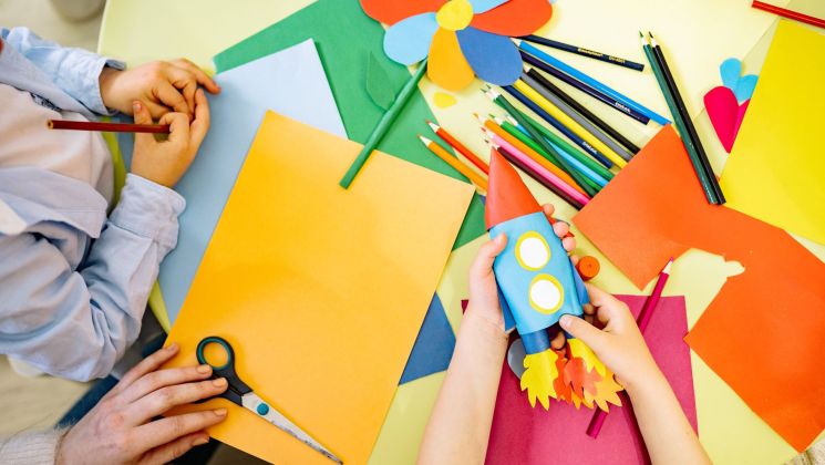 Zwei Kinderhände arbeiten mit buntem Papier, Scheren und Stiften an Bastelprojekten, darunter eine selbstgemachte Rakete.